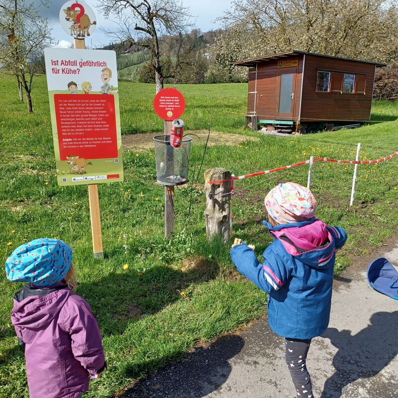I bambini cercano di gettare una lattina di alluminio vuota in un cestino dei rifiuti, accanto al cartello del percorso didattico.
