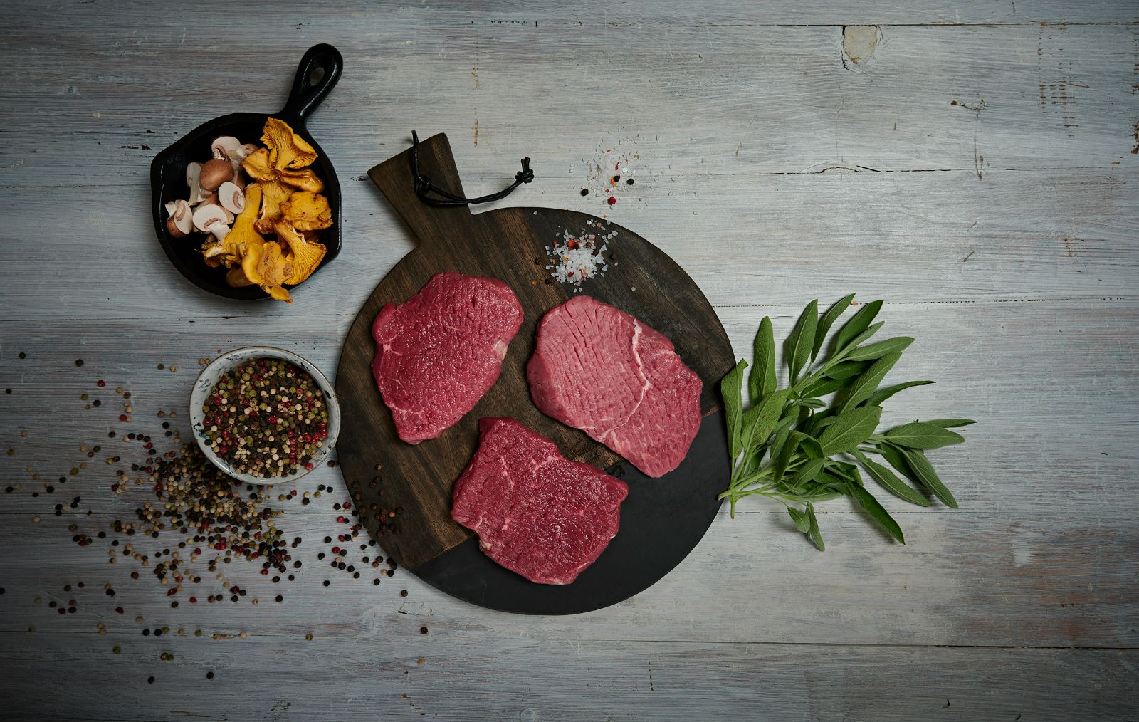 Manzo crudo su una tavola di legno, con accanto sale e pepe in grani, salvia, funghi e finferli